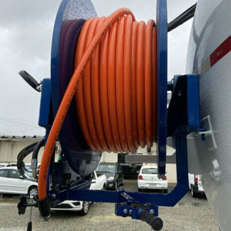 JC7 SEWER JETTING HOSE ASSEMBLY-100MT COILS-2 hydraulicplanet A close-up of a JC7 SEWER JETTING HOSE ASSEMBLY-100M COILS, orange and coiled on a blue metal reel mounted to a vehicle, with parked cars and buildings in the background.